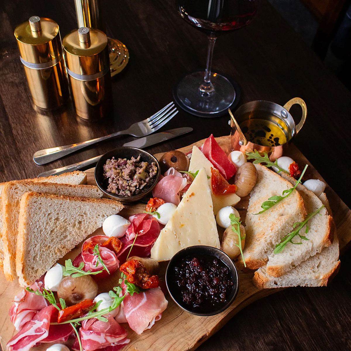 A wooden sharing board with cured ham, cheese, bread, olives and chutney